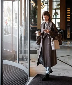 Woman in a long brown coat checking her phone while entering a modern building through a revolving elevator glass door.