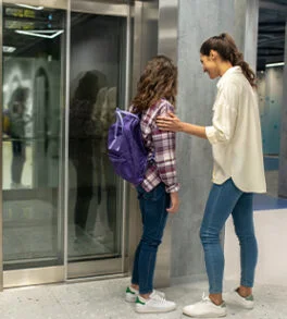 Mother comforting her daughter with a purple backpack as the girl prepares to enter a modern glass elevator.
