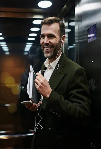 Smiling, stylish businessman in a modern metal elevator wearing earbuds and holding a phone and notebook.